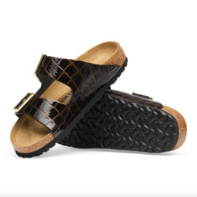 A pair of black crocodile-embossed slide sandals with gold-tone metal buckles and a natural cork footbed and outsole edge