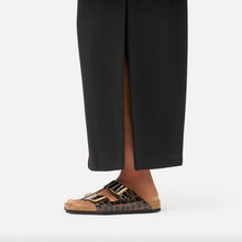 A person wearing a brown and black crocodile-embossed slide sandal with gold-tone buckle, styled with black wide-leg trousers