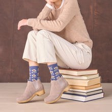 A person seated on a stack of books wearing beige knit pants, a beige sweater, styled in taupe suede Chelsea ankle boots 