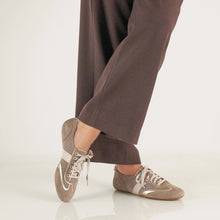 A person wearing brown wide-leg trousers and taupe low-top sneakers with white laces and white stripe accents