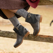 A person styled with black burnished calf ankle boots with brown soles, gray knit socks, and a burgundy skirt