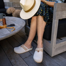Person sitting outdoors wearing white clogs with clork velcro straps, a straw hat, and a patterned dress.