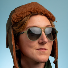 Person wearing a brown aviator hat and sunglasses against a blue background