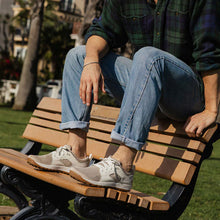 Person sitting on a wooden bench wearing jeans and sneakers in an outdoor setting