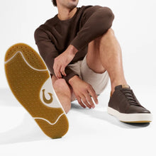 A man wearing a brown sweatshirt and beige shorts, showing off a brown leather low-top sneaker. One shoe is shown in a close-up from the sole, highlighting the textured brown outsole, while the other shoe is shown from the side.