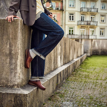A person wearing dark blue jeans, brown leather clogs, and a brown blazer