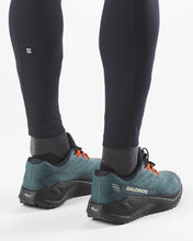 Salomon trail running shoes in teal and black with orange lace hoops, shown on a model wearing black Salomon leggings 